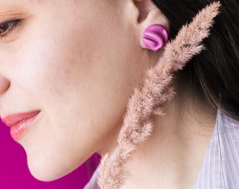 May include: A close-up of a person wearing a pair of bright pink, round earrings. The earrings have a swirled design. A sprig of dried, light pink grass is in the foreground. The background is a solid, bright pink.