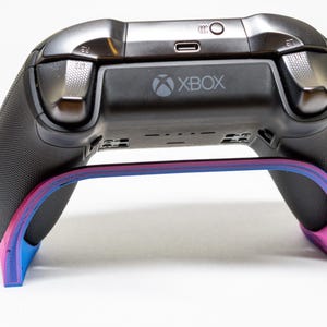 May include: A black Xbox controller rests on a stand with a gradient of pink, blue, and purple. The controller has the Xbox logo and silver accents. The stand is designed to hold the controller.