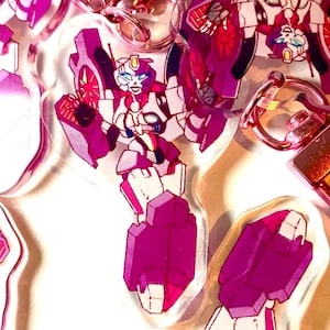 Peut inclure: Une collection de porte-clés en acrylique représentant un personnage robotique dans des tons magenta, blanc et violet. Chaque porte-clés est fixé à un fermoir mousqueton rose métallique. Le design du robot présente une forme humanoïde.