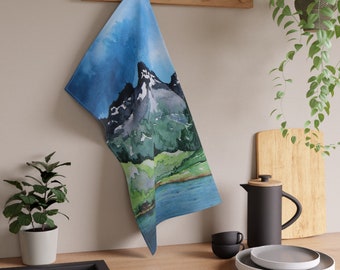 May include: A blue and green kitchen towel with a watercolor painting of mountains and a lake. The towel is hanging on a wooden rack.