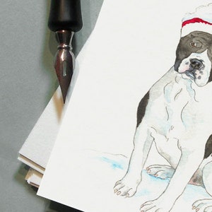 K&ouml;nnte beinhalten: Aquarellmalerei eines schwarz-wei&szlig;en Hundes mit Weihnachtsm&uuml;tze. Der Hund sitzt mit leicht geneigtem Kopf. Eine schwarze Feder ist in der oberen linken Ecke, mit einem Stapel wei&szlig;em Papier.
