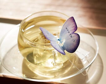 May include: A clear glass teacup filled with yellow tea, resting on a matching saucer. A light purple butterfly-shaped tea infuser is perched on the rim of the cup, with text on the butterfly's wing. The tea is steeping.