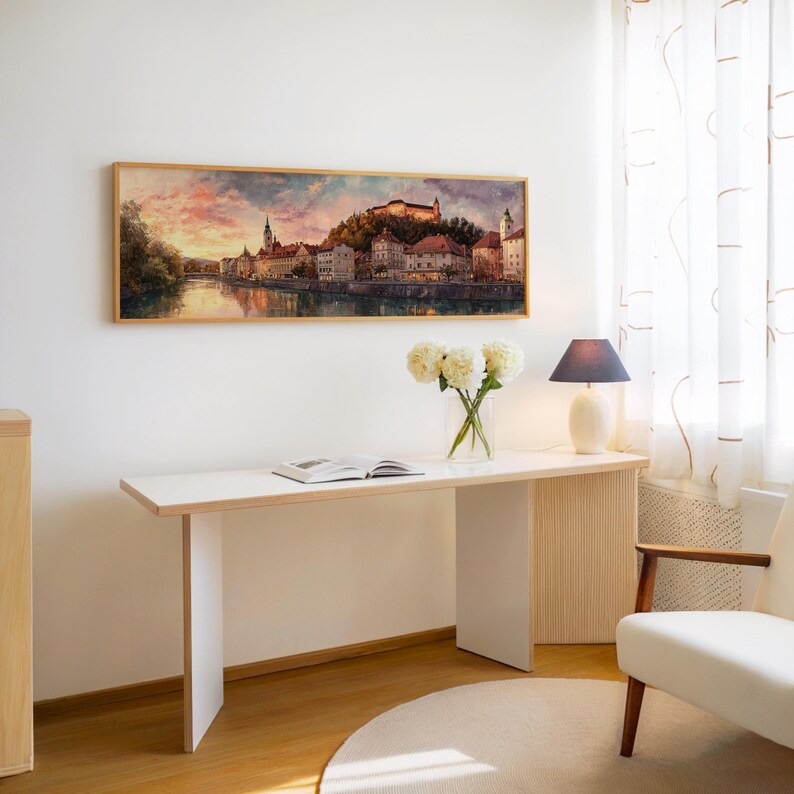 May include: A rectangular framed watercolor painting of a European cityscape hangs above a white desk. A vase of white flowers, a lamp, and an open book are on the desk. A white chair and a round rug are also in the room.
