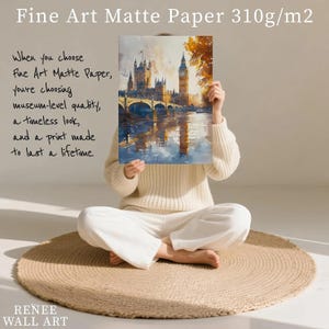 A person is sitting on a round rug, holding up a framed painting of a cityscape with a castle in the background.