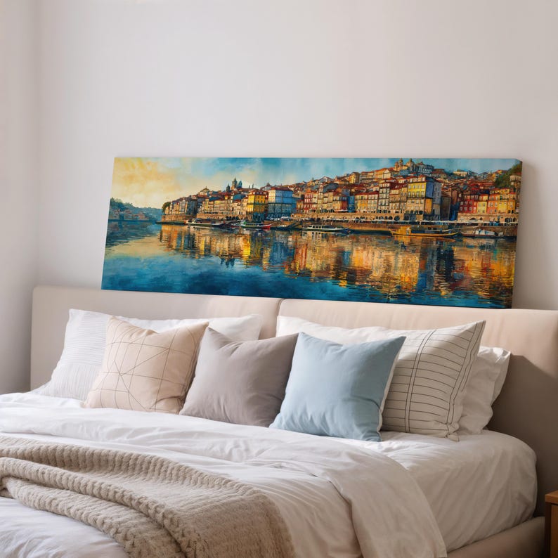 May include: A panoramic canvas print depicting a colorful cityscape reflected in the water. The artwork features buildings in shades of yellow, orange, and red, with a blue sky and water. The print is displayed above a bed with pillows.