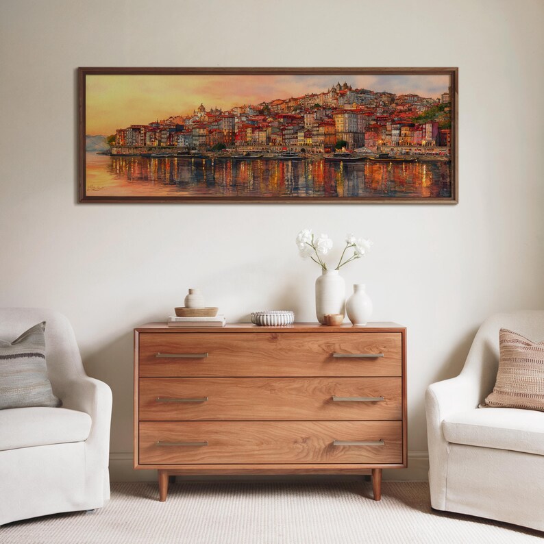 May include: A framed landscape painting of a vibrant cityscape reflected in water hangs above a wooden chest of drawers. Two white armchairs sit either side of the chest, creating a comfortable living space.