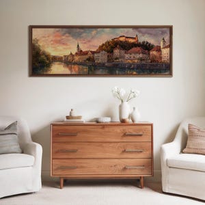 May include: A framed landscape painting depicts a European city at sunset, with a river reflecting the colorful sky. Below, a wooden dresser with three drawers and brass handles is adorned with vases and decorative items. Two white armchairs flank the dresser.