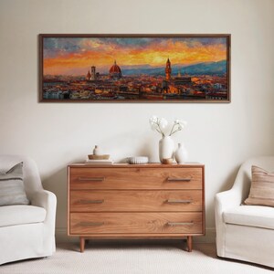 Pu&ograve; includere: Un dipinto incorniciato di un paesaggio urbano al tramonto, con una cupola e una torre prominenti. L'opera d'arte &egrave; appesa sopra un com&ograve; in legno con tre cassetti. Due poltrone bianche affiancano il com&ograve;, creando un'atmosfera da soggiorno.