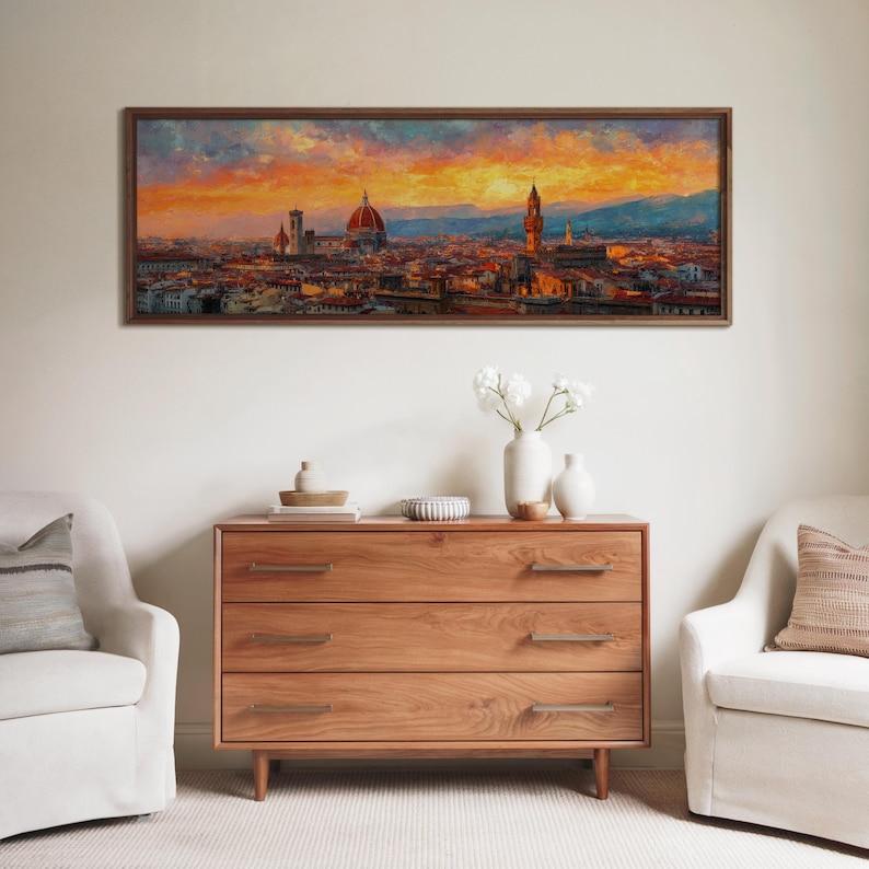 Pu&ograve; includere: Un dipinto incorniciato di un paesaggio urbano al tramonto, con una cupola e una torre prominenti. L'opera d'arte &egrave; appesa sopra un com&ograve; in legno con tre cassetti. Due poltrone bianche affiancano il com&ograve;, creando un'atmosfera da soggiorno.