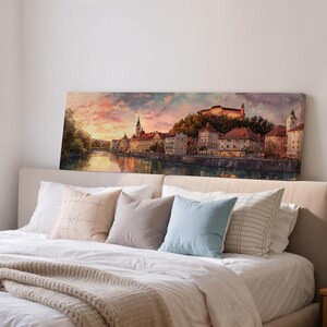 May include: A long rectangular canvas print depicting a European cityscape at sunset. The artwork features buildings along a river, a castle on a hill, and a colorful sky with pink and orange hues. The print is displayed above a bed with pillows.