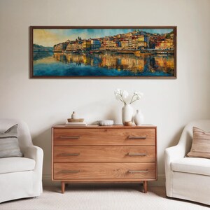 May include: A framed watercolor painting of a cityscape with colorful buildings reflected in the water. The painting hangs above a wooden dresser with three drawers. Two white armchairs flank the dresser, creating a cozy living room scene.
