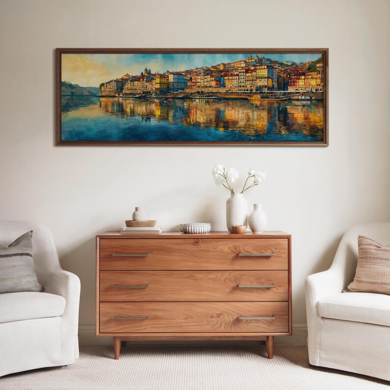 May include: A framed watercolor painting of a cityscape with colorful buildings reflected in the water. The painting hangs above a wooden dresser with three drawers. Two white armchairs flank the dresser, creating a cozy living room scene.