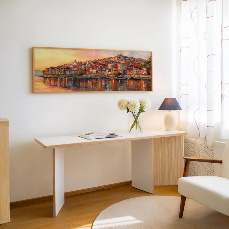 May include: A long, rectangular framed painting of a cityscape at sunset, with buildings reflected in the water. A white desk with a book, flowers, and a lamp sits below the painting. A white chair is to the right.