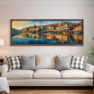 May include: A framed watercolor painting depicts a colorful cityscape reflected in the water. The artwork features buildings in shades of yellow, orange, and red, with a dark brown frame. The painting hangs above a white sofa with gray and checkered pillows.