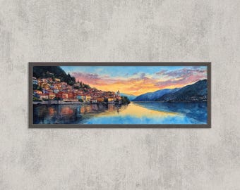 May include: A framed landscape painting depicts a lakeside village at sunset. The artwork features colorful buildings nestled on a hillside, reflecting in the calm water. The sky transitions from orange to blue, with mountains in the background.