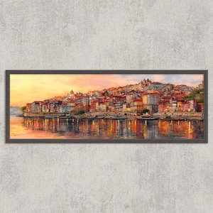 May include: A framed watercolour painting of a cityscape at sunset. The artwork depicts buildings with red tile roofs along a river, reflecting in the water. The sky transitions from yellow to orange, creating a warm, inviting scene.