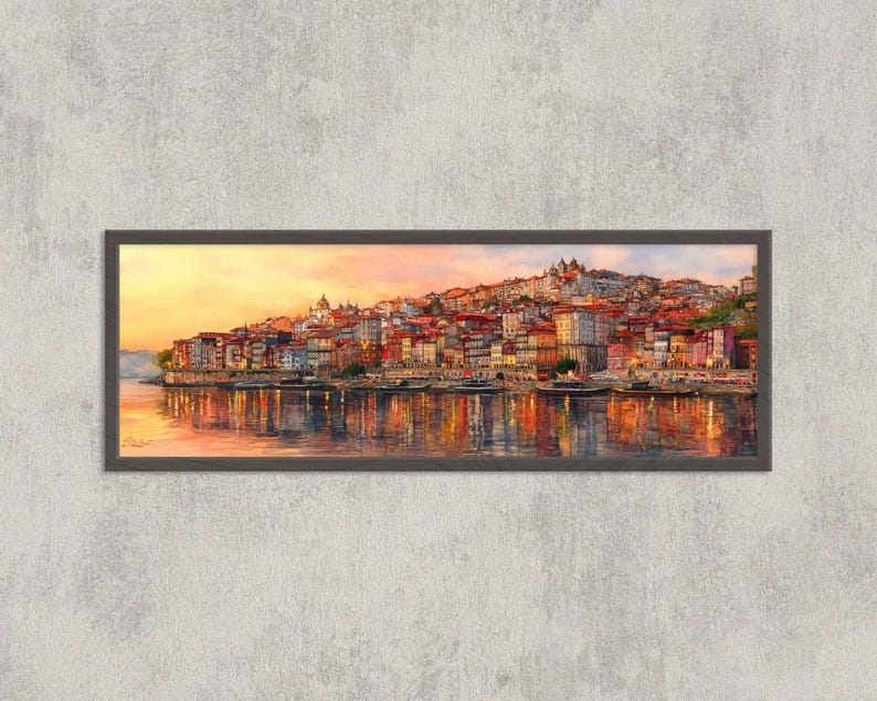 May include: A framed watercolour painting of a cityscape at sunset. The artwork depicts buildings with red tile roofs along a river, reflecting in the water. The sky transitions from yellow to orange, creating a warm, inviting scene.