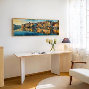 May include: A watercolor painting of a cityscape with buildings reflected in the water, framed in a light wood. The painting hangs above a white desk with a vase of white flowers, a lamp, and an open book. A white chair is to the right.