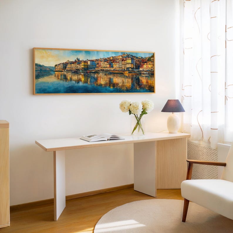 May include: A watercolor painting of a cityscape with buildings reflected in the water, framed in a light wood. The painting hangs above a white desk with a vase of white flowers, a lamp, and an open book. A white chair is to the right.