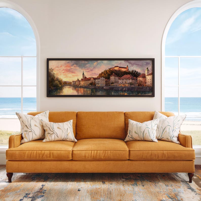 May include: A framed landscape painting of a European city with a river, buildings, and a castle. The painting hangs above a mustard-colored sofa with decorative pillows. The room has large arched windows overlooking the ocean.