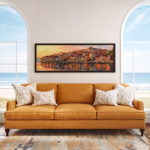 May include: A three-seater mustard sofa with decorative pillows. Above the sofa is a framed landscape painting of a waterside city. The room has large arched windows overlooking the sea.