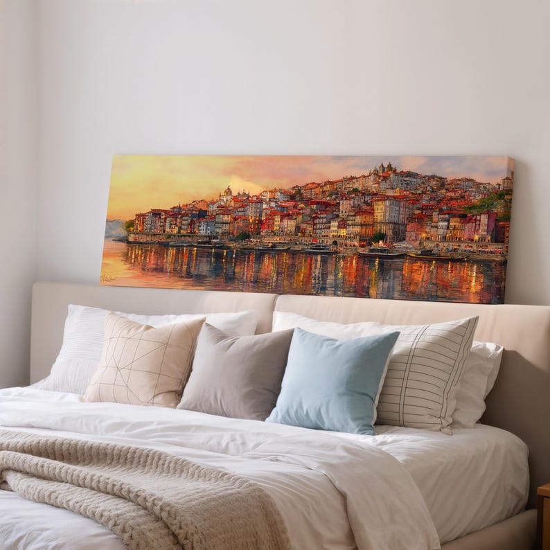 May include: A long canvas print of a waterside cityscape at sunset. The artwork shows buildings in red, orange, and yellow, reflected in the water. The print is displayed above a bed with pillows and a blanket, creating a warm, inviting atmosphere.