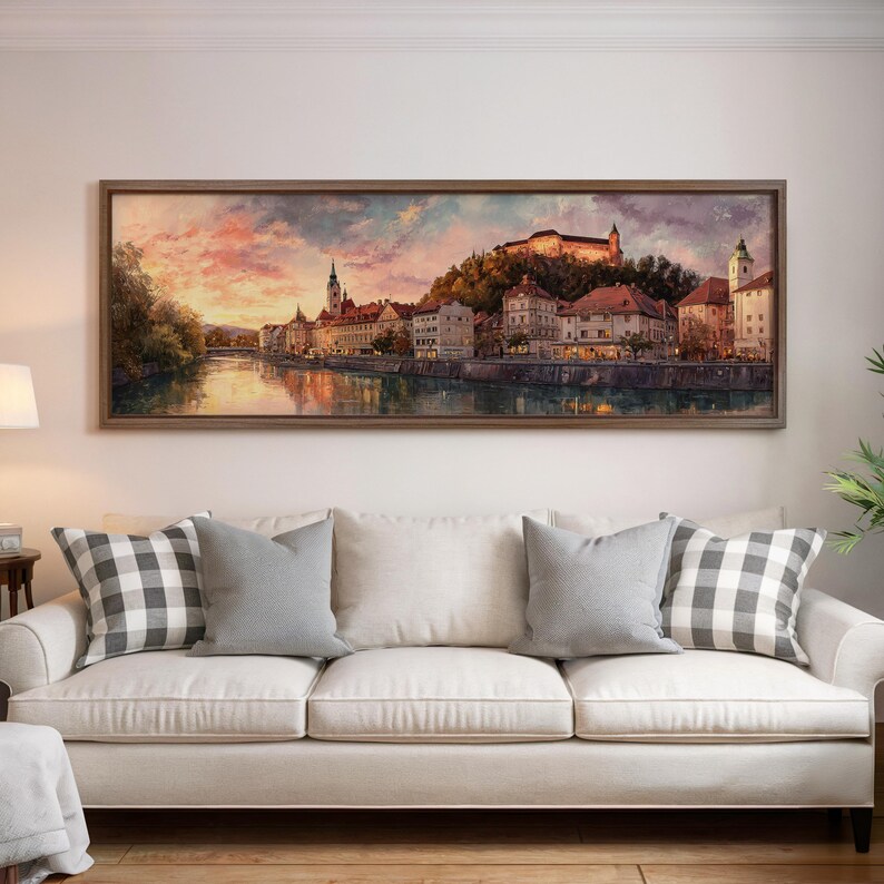 May include: A framed landscape painting depicts a European city at sunset. The artwork showcases buildings along a river, with a castle on a hill. The sky is a blend of pink, orange, and purple hues. The painting is displayed above a white sofa with gray pillows.