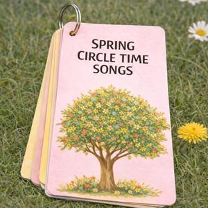 May include: A stack of colorful cards held together by a metal ring. The top card is pink and features the text "SPRING CIRCLE TIME SONGS" above a tree illustration with colorful flowers. The cards are resting on green grass with daisies.