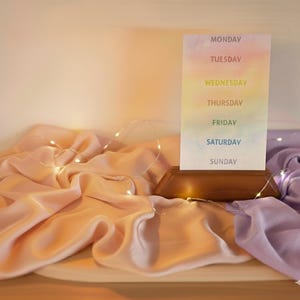 May include: A weekly planner card with a gradient background of pastel colors, held in a wooden base. The days of the week are listed vertically. Soft pink and purple fabric is draped around the base, with string lights.
