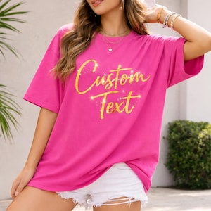 May include: A bright pink t-shirt with short sleeves and the words "Custom Text" in gold glitter script. The shirt is a loose fit and appears to be made of a soft material. The model is wearing white shorts.