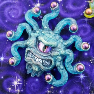May include: A blue and green resin figurine of a beholder with a single large eye and multiple tentacles. The eye has a purple and pink iridescent finish. The tentacles have iridescent purple spheres at the ends.