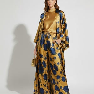 May include: A gold satin ensemble featuring a high-neck top, wide-leg pants, and a long kimono-style jacket. The outfit is adorned with a large blue floral pattern. A small gold handbag and gold heels complete the look.