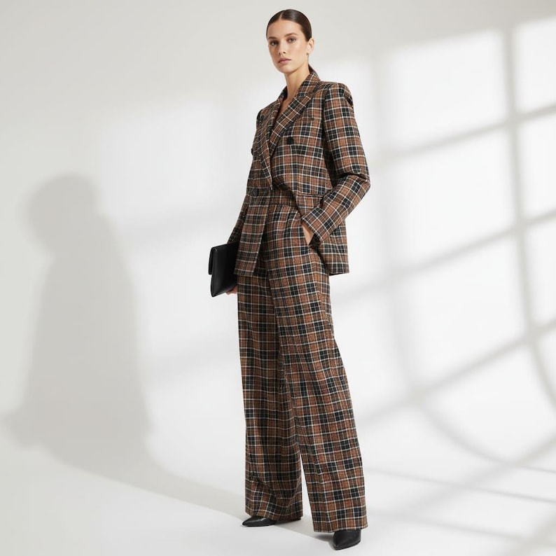 May include: A brown, black, and white plaid suit, consisting of a double-breasted blazer and wide-leg trousers. The model is holding a black clutch. The suit is a classic, tailored design.