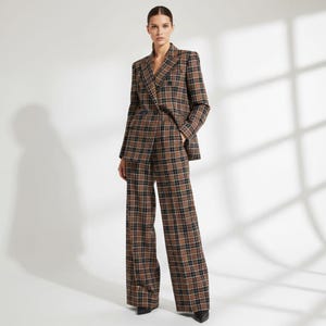 May include: A brown, black, and tan plaid suit, consisting of a double-breasted blazer and wide-leg trousers. The suit is worn by a person standing in front of a white background. The blazer has a notched lapel and the trousers are high-waisted.