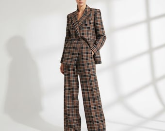 May include: A brown, black, and tan plaid suit, consisting of a double-breasted blazer and wide-leg trousers. The suit is worn by a person standing in front of a white background. The blazer has a notched lapel and the trousers are high-waisted.