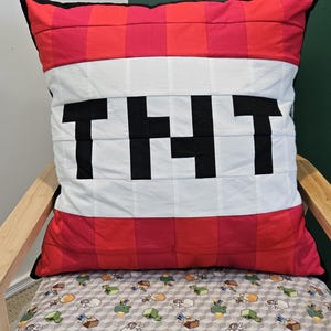 May include: A square cushion with a TNT block design. The cushion has a red and white colour scheme with the word "TNT" in large black letters. The cushion is placed on a chair with a Minecraft-themed patterned cushion.