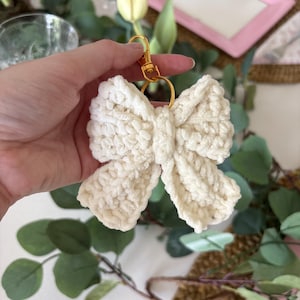 May include: A cream-colored, crocheted bow keychain with a gold-colored clasp and ring. The bow is made of a soft, textured yarn. The background includes greenery and a pink plate.