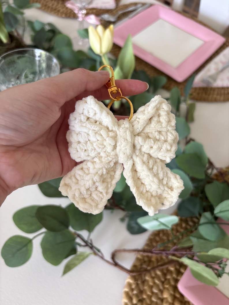 May include: A cream-colored, crocheted bow keychain with a gold-colored clasp and ring. The bow is made of a soft, textured yarn. The background includes greenery and a pink plate.