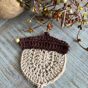 May include: A handmade crochet acorn in shades of brown and beige. The top is a dark brown, and the body is a lighter beige. The acorn is placed on a light blue wooden surface, with fall-themed decorations in the background.