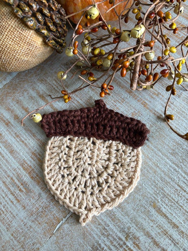 May include: A handmade crochet acorn in shades of brown and beige. The top is a dark brown, and the body is a lighter beige. The acorn is placed on a light blue wooden surface, with fall-themed decorations in the background.