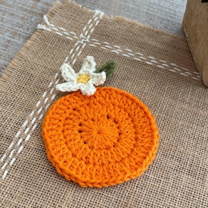 Puede incluir: Un posavasos de ganchillo naranja hecho a mano con forma de rodaja de naranja, con una flor blanca y amarilla y un acento de hoja verde. El posavasos está colocado sobre un mantel individual de arpillera con costuras blancas.