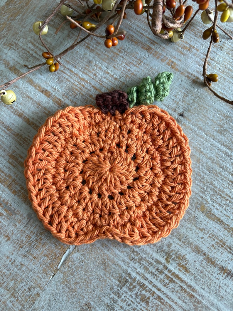 May include: A handmade, orange crochet pumpkin coaster with a brown stem and green leaves. The coaster is round and has a textured appearance, perfect for fall decor. The pumpkin is made of yarn.