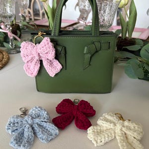 May include: A green handbag with a top handle and a pink crocheted bow keychain attached. Additional crocheted bow keychains in blue, red, and cream are displayed below the bag. The bows have gold-colored clasps.