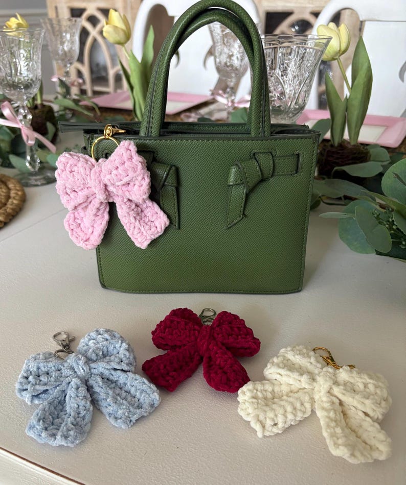 May include: A green handbag with a top handle and a pink crocheted bow keychain attached. Additional crocheted bow keychains in blue, red, and cream are displayed below the bag. The bows have gold-colored clasps.