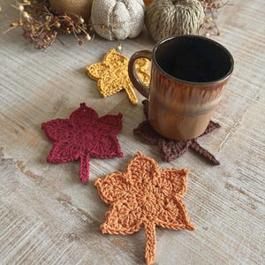 以下が含まれることがあります： 茶色のセラミックマグカップが、茶色の葉の形をしたかぎ針編みのコースターの上に置かれています。赤、黄、オレンジ色の葉のコースターが3つあります。コースターは明るい色の木製の表面に置かれています。
