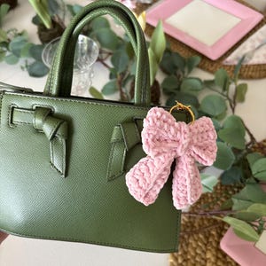 May include: A green handbag with a textured finish, featuring a bow detail on the front and a matching handle. A pink crocheted bow charm hangs from the handle. The bag is set against a table with floral decorations and pink plates.