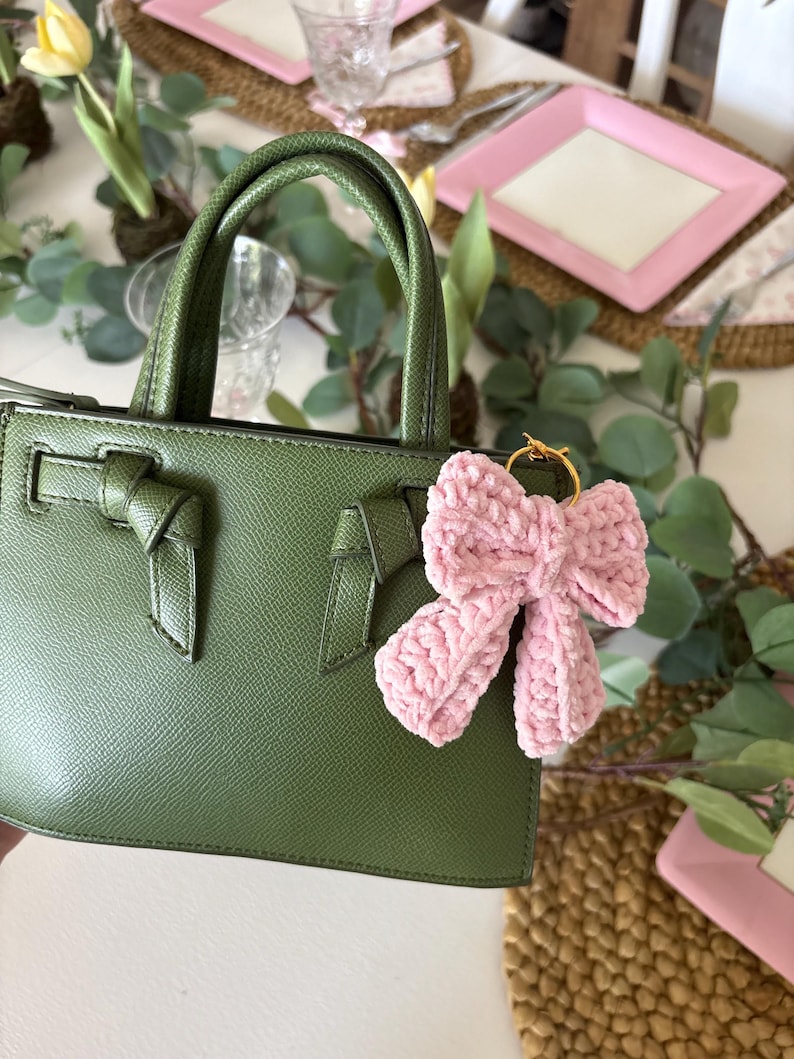 May include: A green handbag with a textured finish, featuring a bow detail on the front and a matching handle. A pink crocheted bow charm hangs from the handle. The bag is set against a table with floral decorations and pink plates.