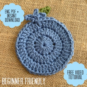 May include: A handmade, blue crochet coaster shaped like a blueberry, with a green leaf detail. The coaster is on a burlap background. Text on the image reads "ENG PDF + INSANT DOWNLOAD", "BEGINNER FRIENDLY", and "FREE VIDEO TUTORIAL".