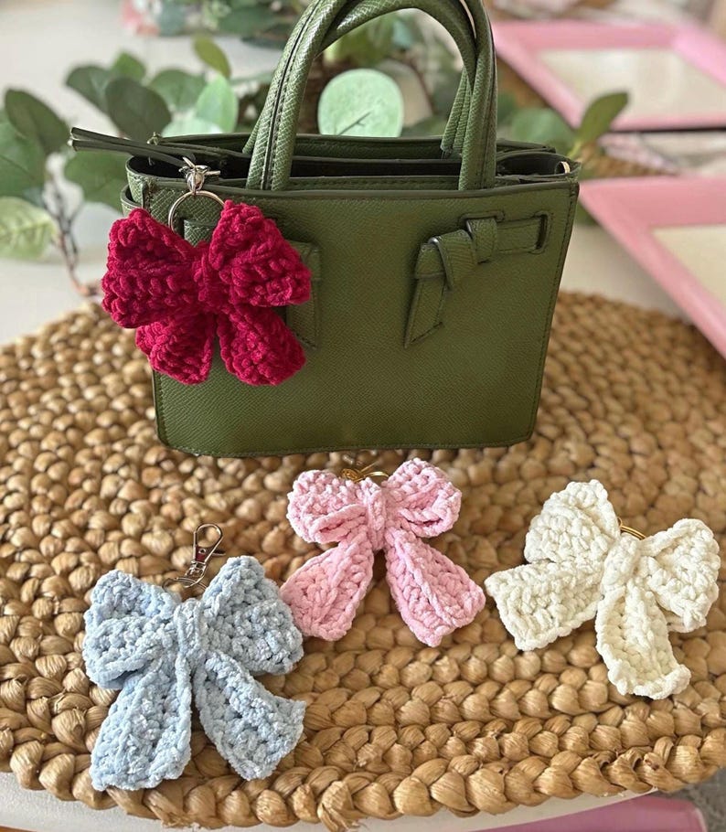 May include: A small, olive green handbag with a red crocheted bow keychain attached. Three additional crocheted bow keychains in light blue, pink, and white are displayed on a woven mat. The bows are handmade and fluffy.
