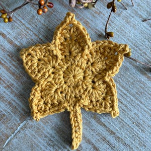 May include: A handmade, golden yellow crochet leaf. The leaf is detailed with visible stitches and a textured surface. The leaf is placed on a light blue wooden surface, with fall-themed decorative branches in the background.